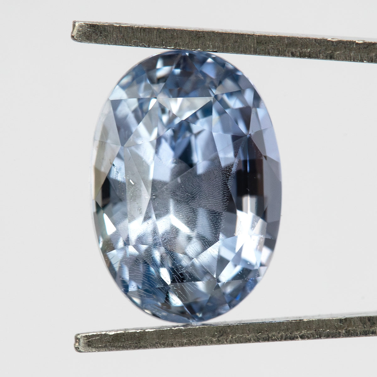 Face-up light blue oval sapphire held vertically between tweezers, showing the outline, pastel color and reflective facets.