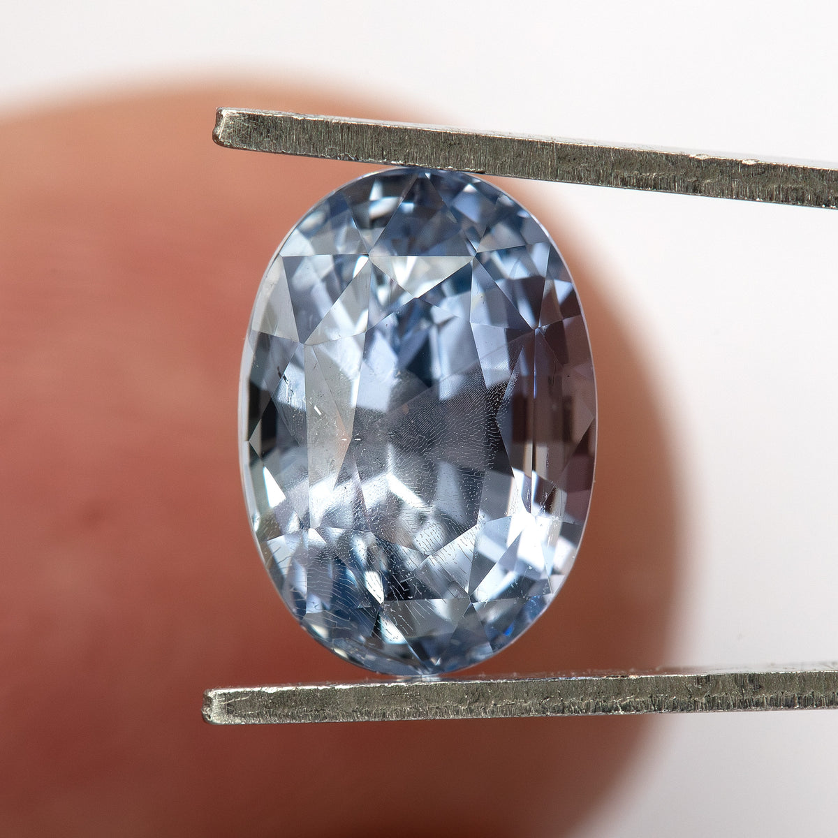 Face-up sapphire between tweezers in front of a fingertip, showing its oval proportions and blue reflections.