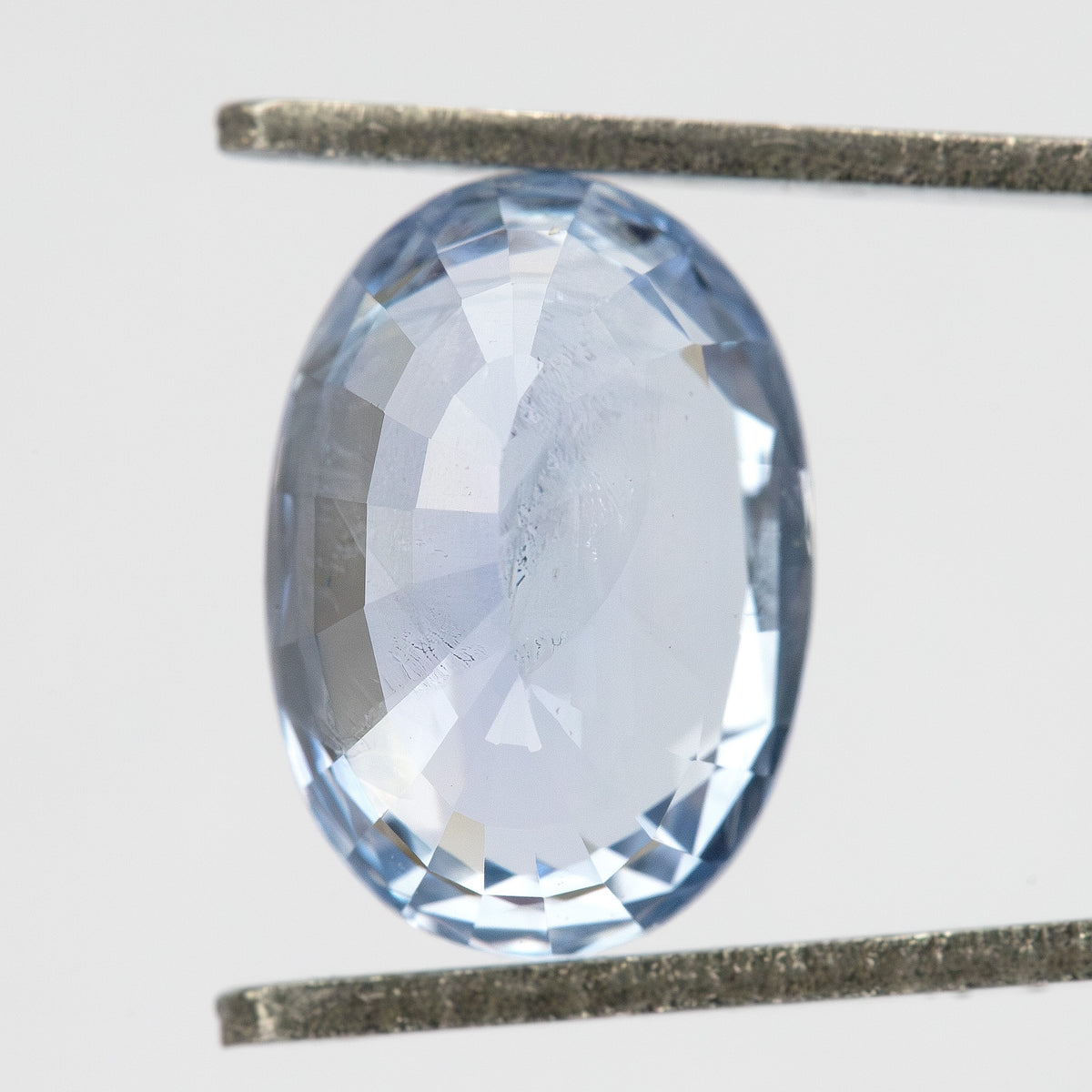 Pavilion-facing view of the oval sapphire held vertically between tweezers, showing its back facets and pale blue color.
