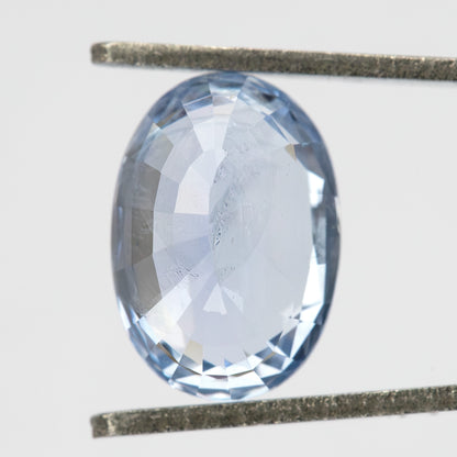 Pavilion-facing view of the oval sapphire held vertically between tweezers, showing its back facets and pale blue color.