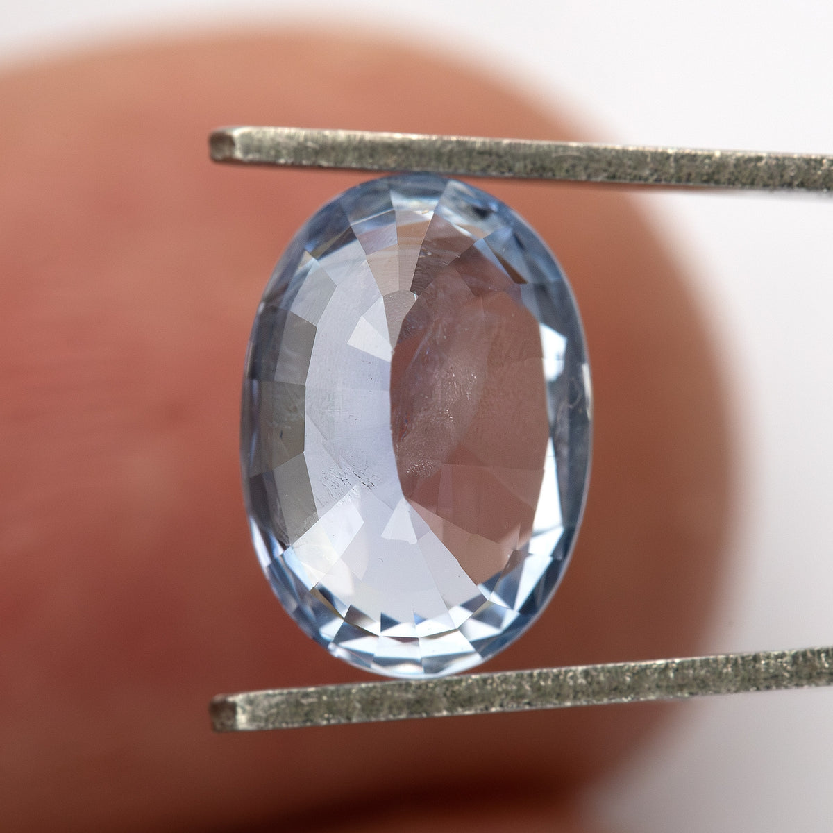 Pavilion-facing oval sapphire between tweezers with a fingertip behind it, showing its back facets and approximate scale.
