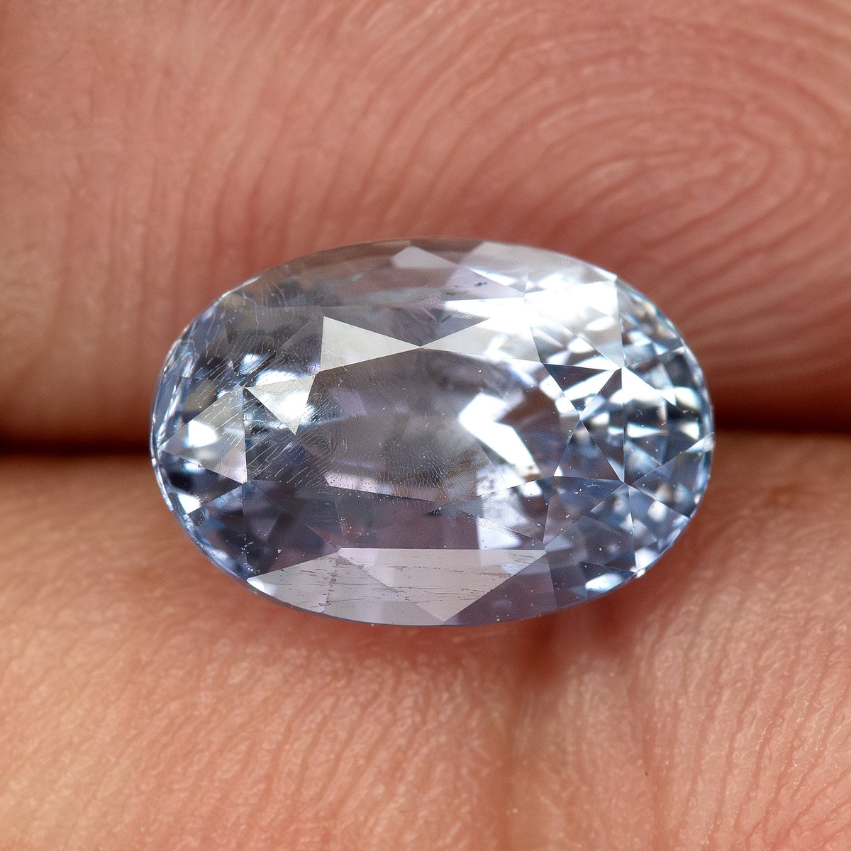 Face-up oval sapphire resting on a fingertip for scale, showing its light blue color and natural internal features.