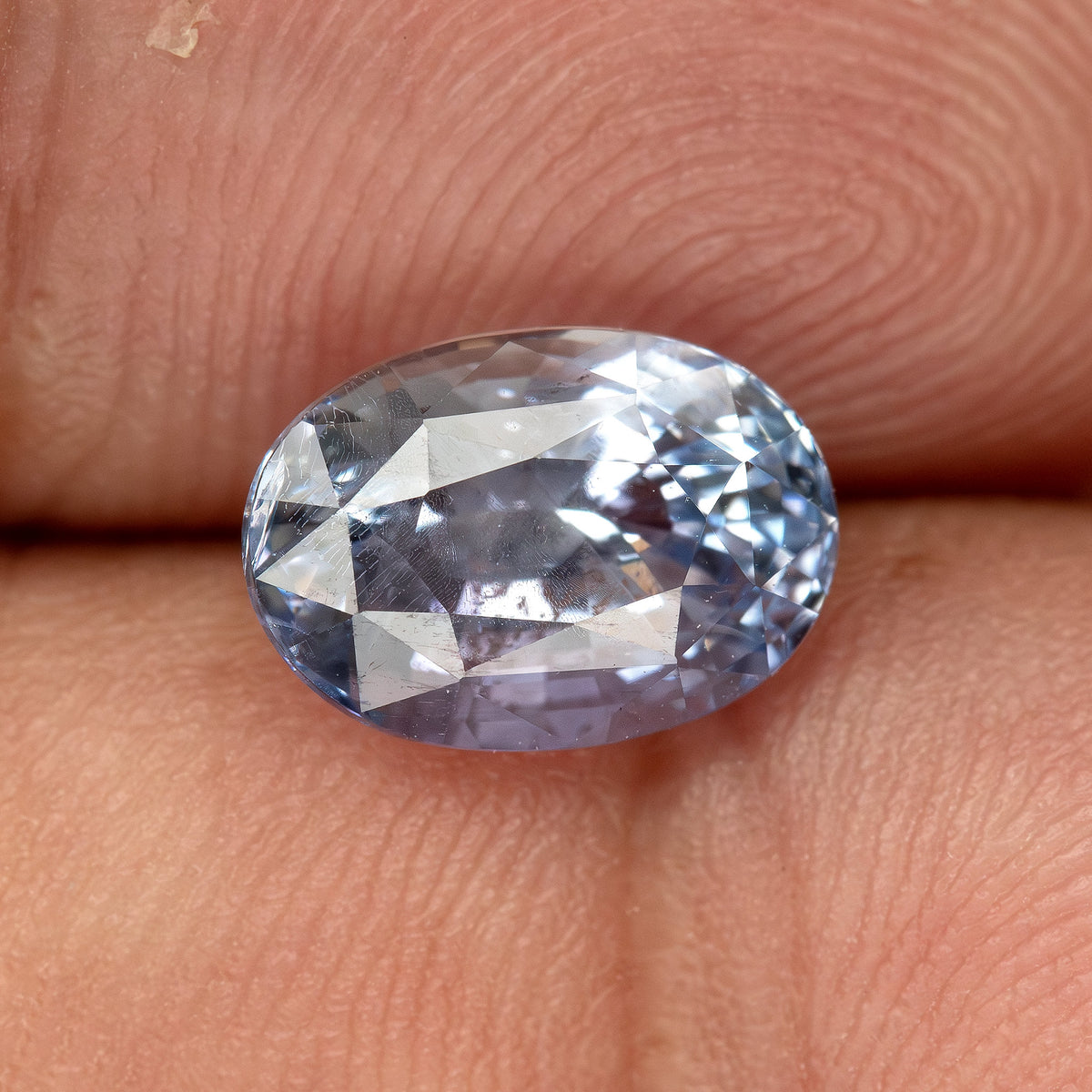 Angled oval sapphire on a fingertip for scale, showing pale blue color, reflections and natural internal features.