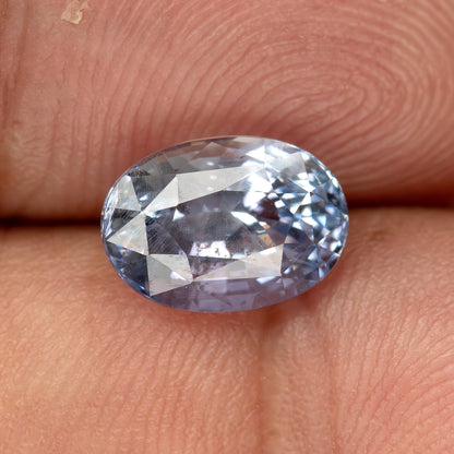 Angled oval sapphire on a fingertip for scale, showing pale blue color, reflections and natural internal features.