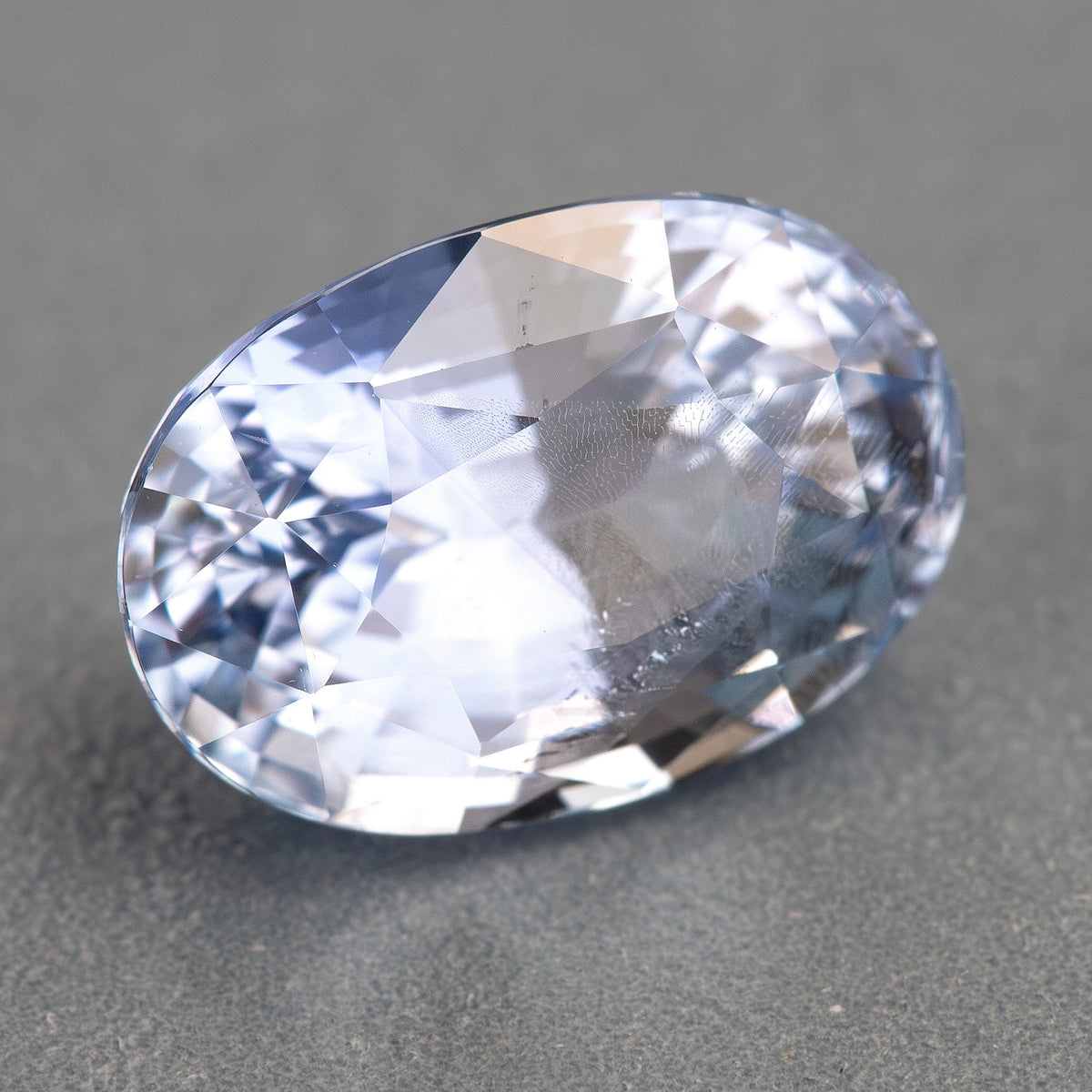 Face-up sapphire on a gray background, showing a bright central reflection and cooler blue edge facets.