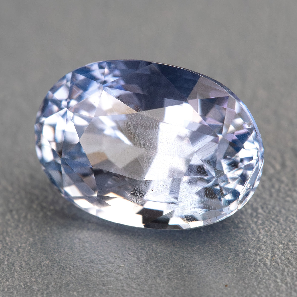 Alternate angled view of the oval sapphire on a gray background, showing pale blue and violet-blue reflections.