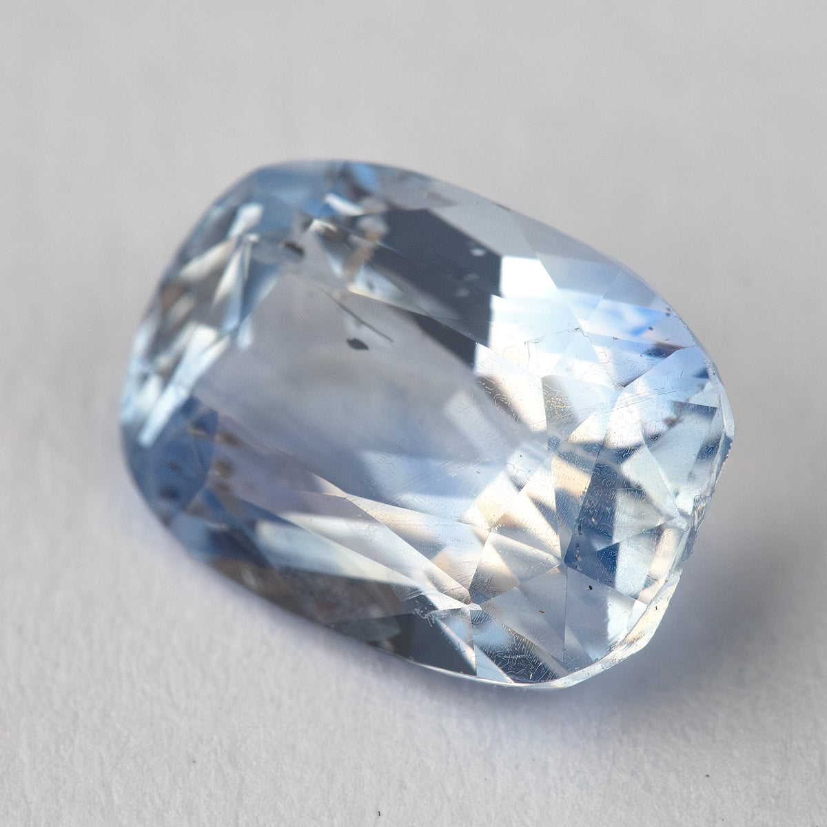 Zafiro de cojín alargado azul claro inclinado sobre blanco, mostrando brillantes reflejos de corona y características internas.