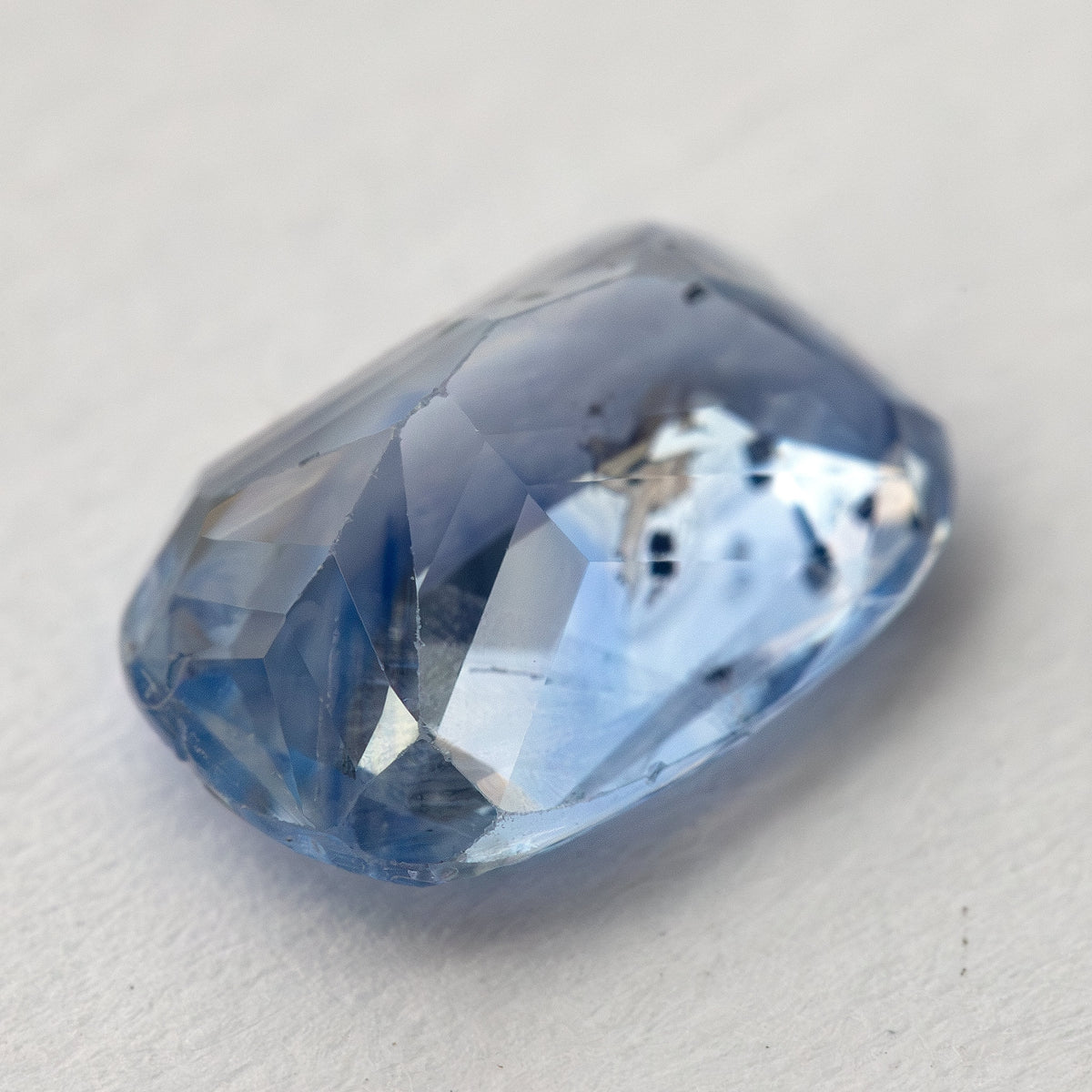 Zafiro de cojín azul claro inclinado hacia el lado del pabellón hacia la cámara sobre blanco, con características internas oscuras visibles.
