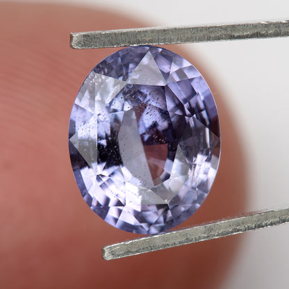 Lavender-blue oval sapphire with visible inclusions held in tweezers against a finger. View 17.