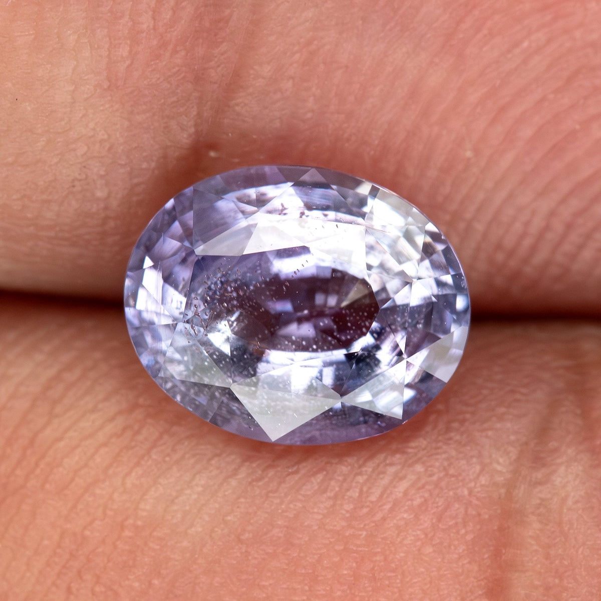Lavender-blue oval sapphire with visible inclusions resting on fingertips for scale. View 3.