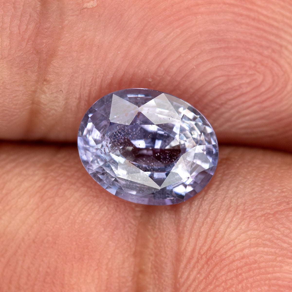 Lavender-blue oval sapphire with visible inclusions resting on fingertips for scale. View 5.
