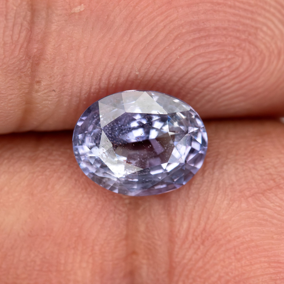 Lavender-blue oval sapphire resting face-up on fingertips, tilted to show pale flashes across its visible inclusions.
