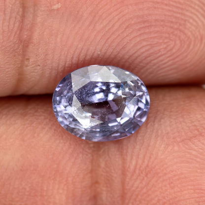 Lavender-blue oval sapphire resting face-up on fingertips, tilted to show pale flashes across its visible inclusions.
