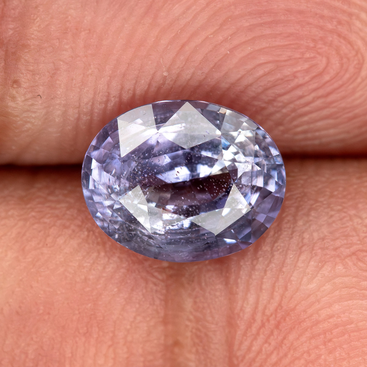 Lavender-blue oval sapphire with visible inclusions resting on fingertips for scale. View 6.