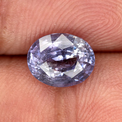 Lavender-blue oval sapphire with visible inclusions resting on fingertips for scale. View 6.