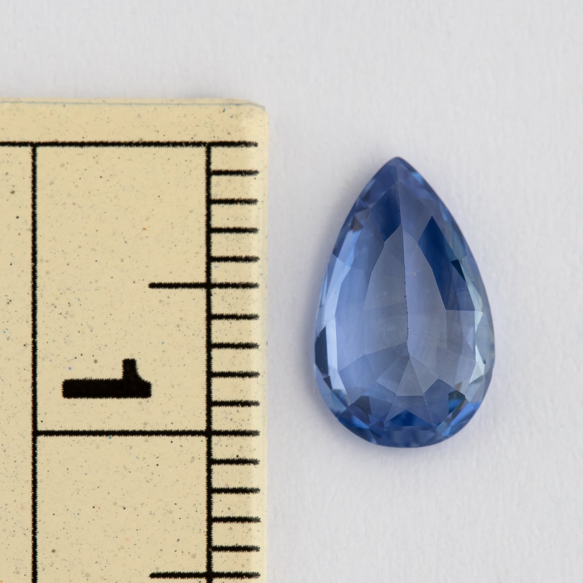Face-up pear mixed-cut sapphire beside a millimeter ruler, showing its approximately 10 mm length, medium violetish blue color and darker blue edge facets.