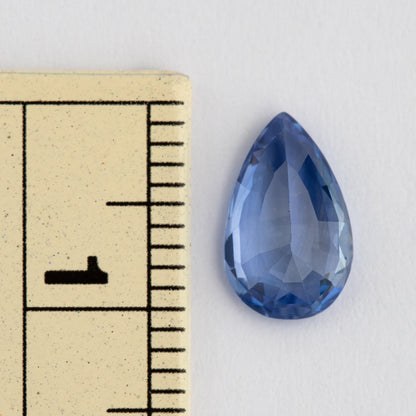 Face-up pear mixed-cut sapphire beside a millimeter ruler, showing its approximately 10 mm length, medium violetish blue color and darker blue edge facets.