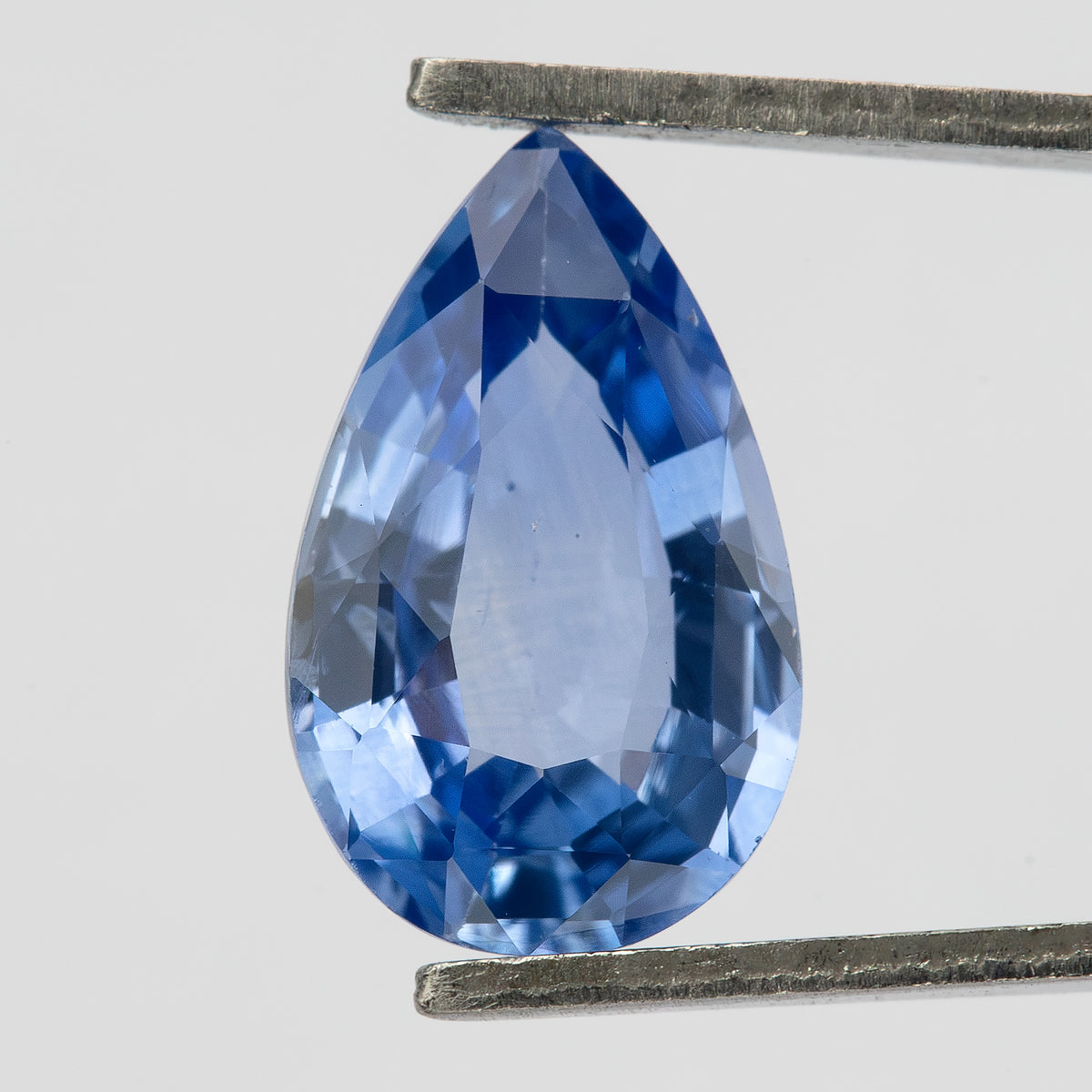 Upright macro view of the pear sapphire held in tweezers, showing its mixed facet pattern, medium-light violet-blue body color and small dark pinpoints under the table.