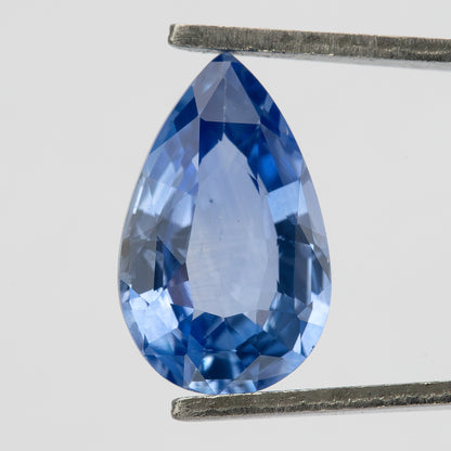 Upright macro view of the pear sapphire held in tweezers, showing its mixed facet pattern, medium-light violet-blue body color and small dark pinpoints under the table.