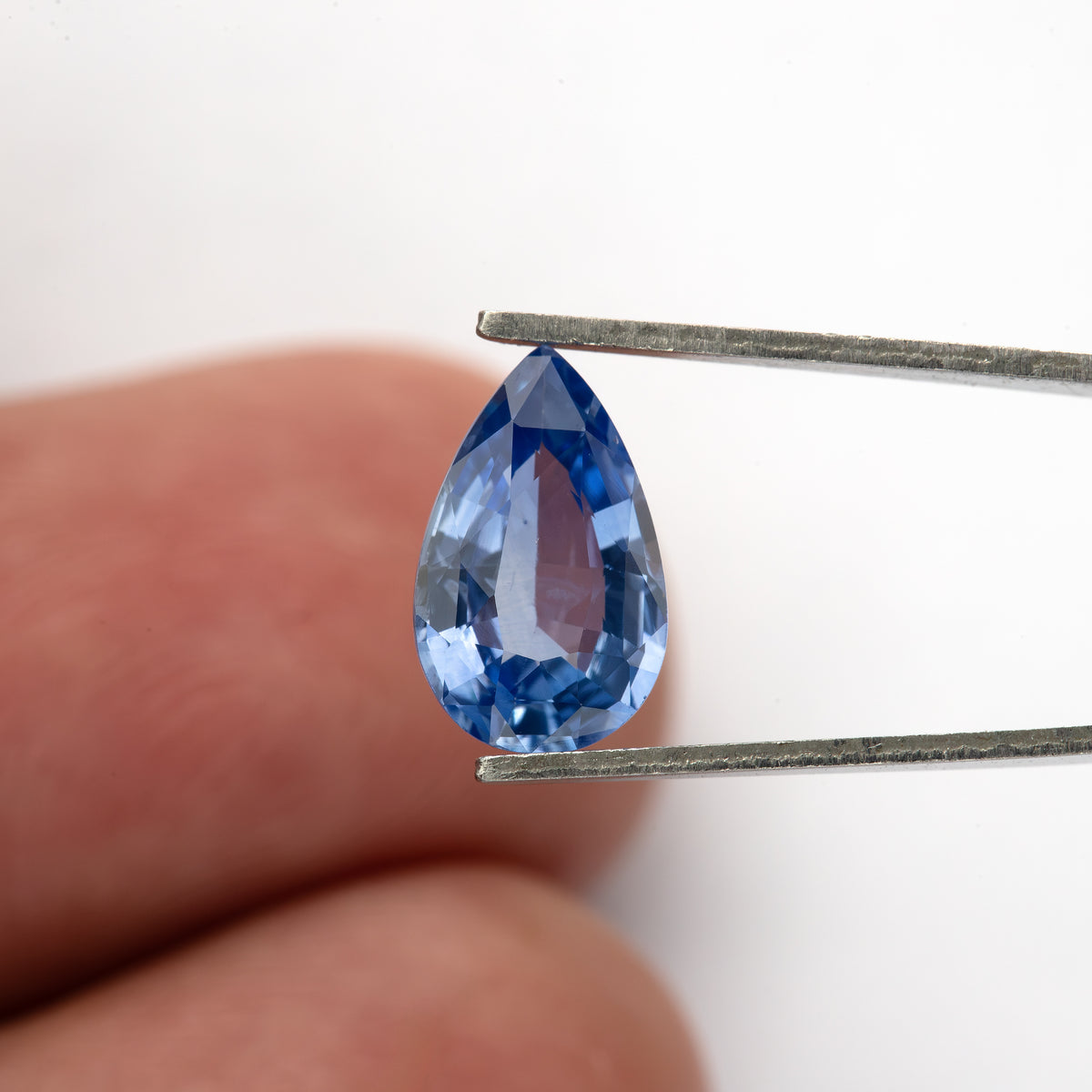 Full pear sapphire held vertically in tweezers in front of fingers for scale, showing a medium-light violet-blue center and vivid darker blue edge facets.