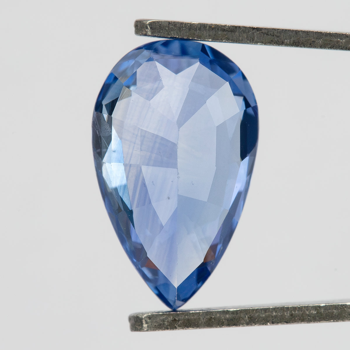 Pavilion-side macro of the pear mixed-cut sapphire in tweezers, showing broad pale violet-blue facets, darker blue edges and the pointed pavilion end.