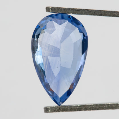 Pavilion-side macro of the pear mixed-cut sapphire in tweezers, showing broad pale violet-blue facets, darker blue edges and the pointed pavilion end.