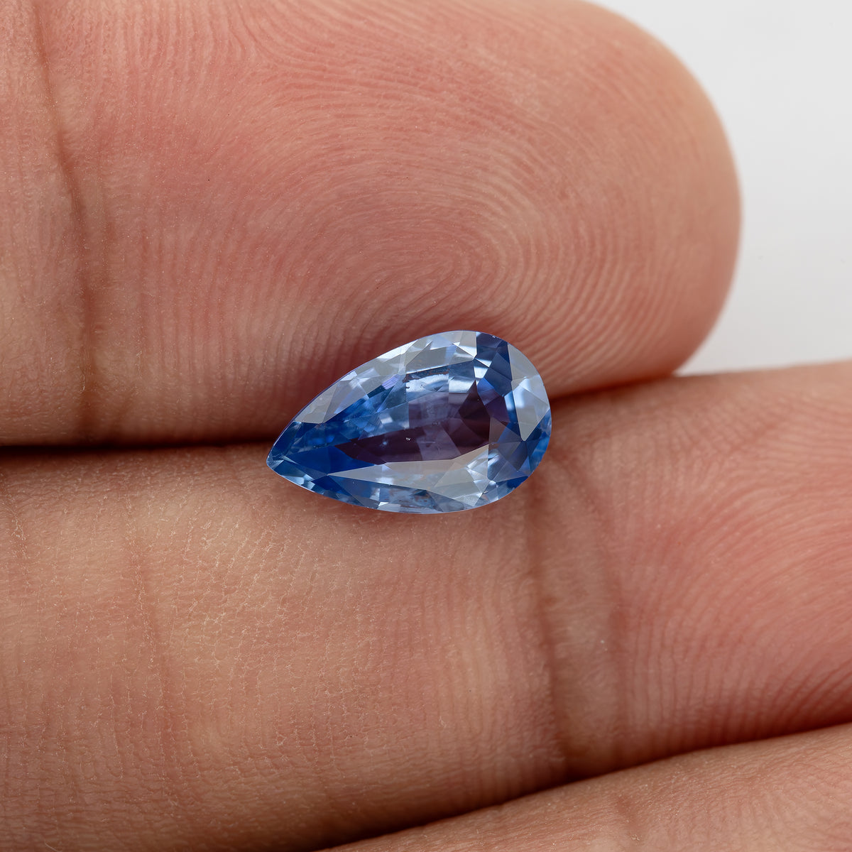 Pear mixed-cut sapphire resting across fingers for scale, with light violet-blue facets, deeper blue edges and a purplish reflection through the center.
