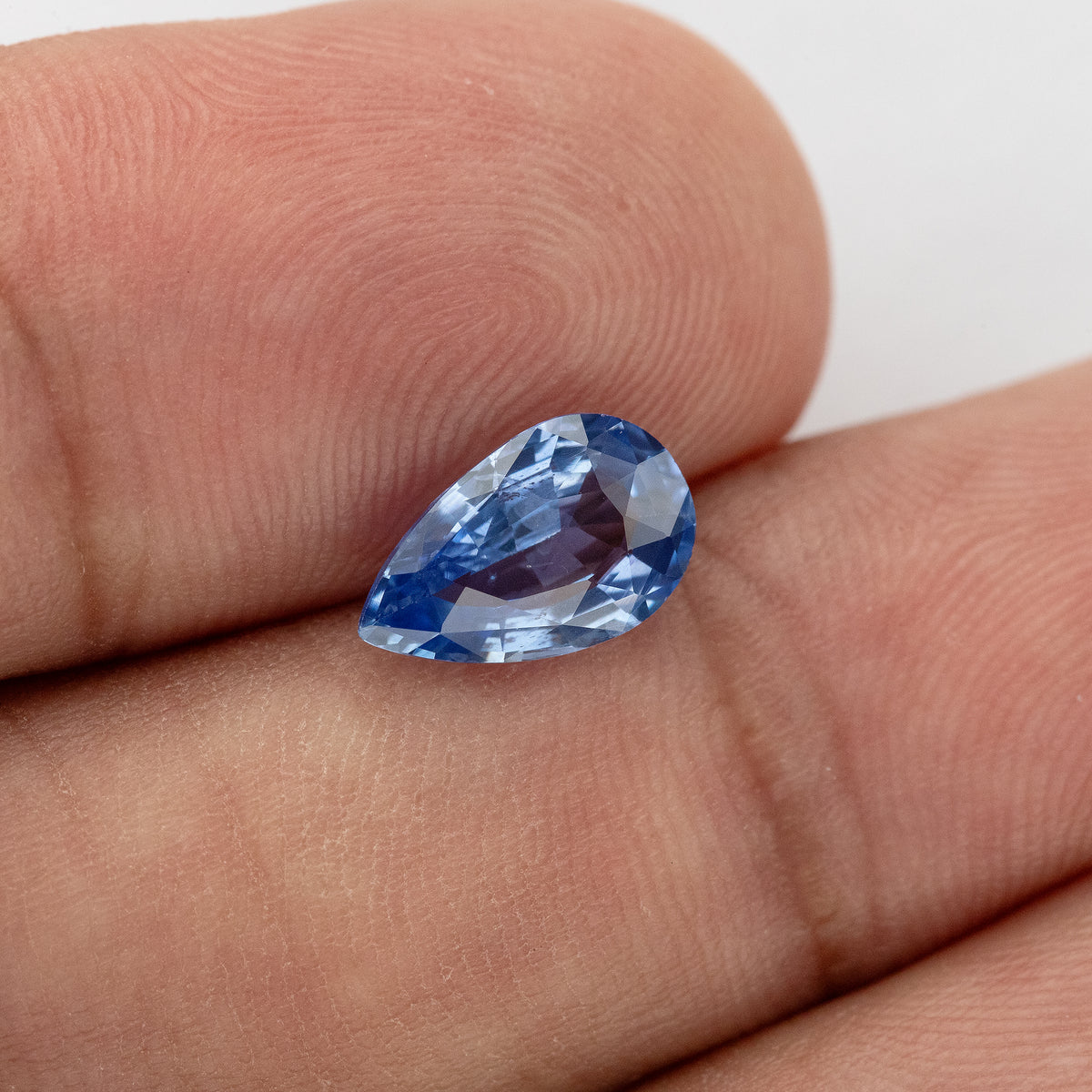 Rotated hand-scale view of the pear sapphire, showing a pale blue crown, dark blue flashes along the edge and a violet reflection from the fingers beneath.