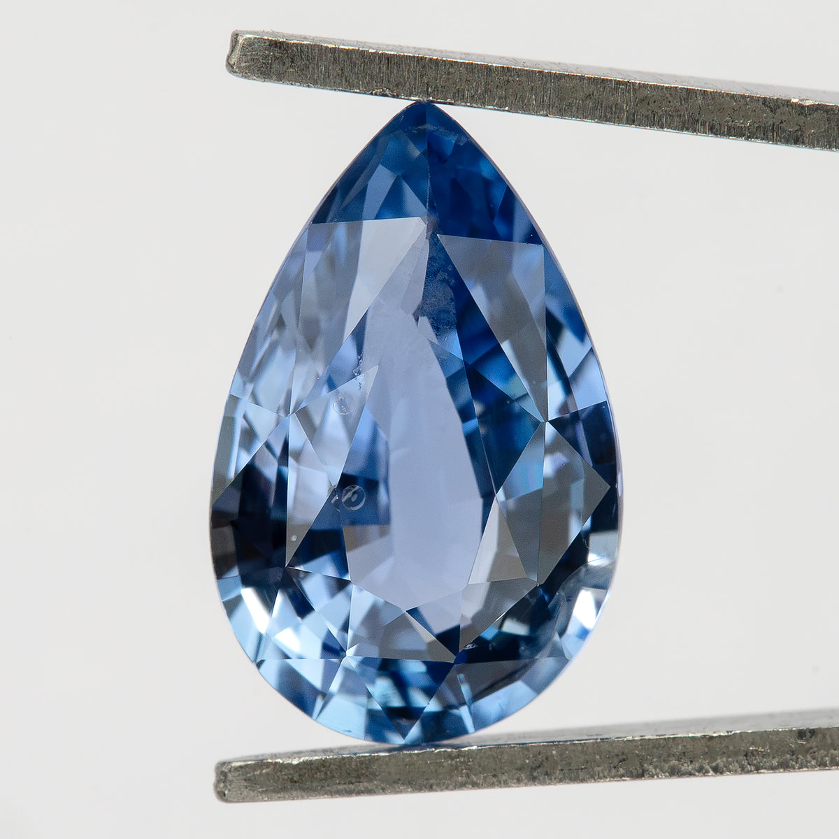 Pear-shaped light blue sapphire held vertically in metal tweezers, showing its face-up outline and mixed-cut facet pattern.