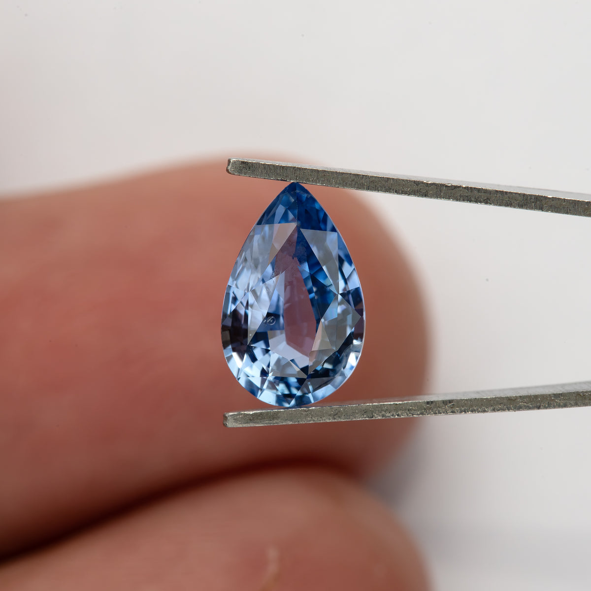 Pear-shaped sapphire held in tweezers in front of a fingertip, showing its real-world size and light blue face-up appearance.