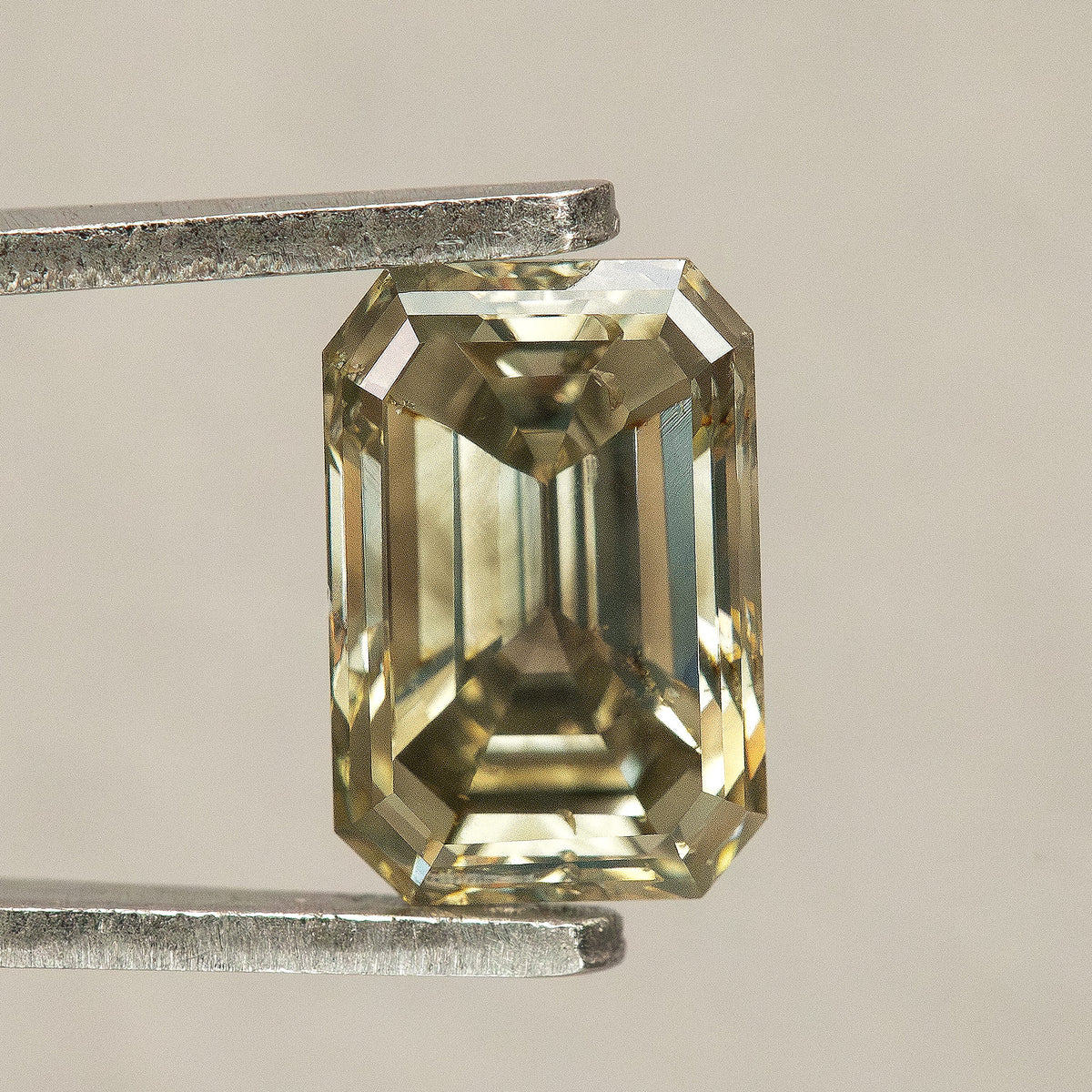 Face-up greenish-brown emerald-cut diamond in tweezers against a pale background, showing clipped corners, step facets and small visible characteristics near the upper-left edge.