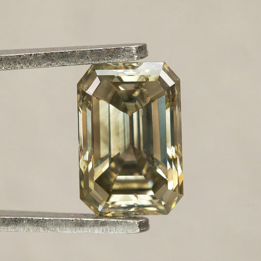 Face-up greenish-brown emerald-cut diamond in tweezers against a pale background, showing clipped corners, step facets and small visible characteristics near the upper-left edge.