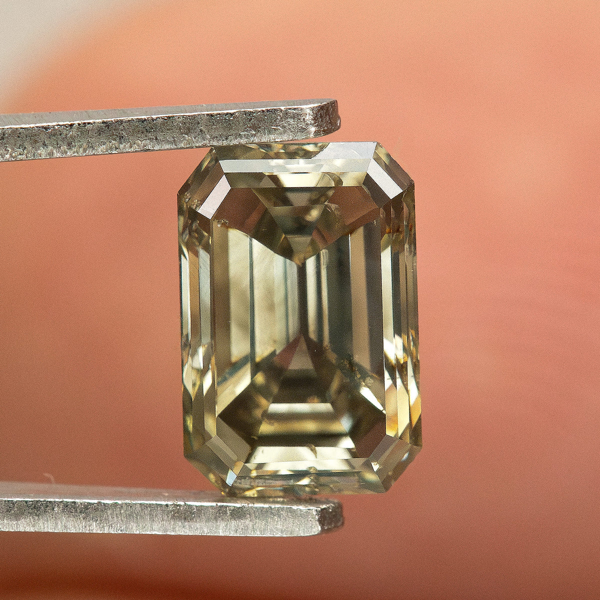 Emerald-cut diamond held upright in tweezers in front of a fingertip, with olive-brown and pale yellow reflections.