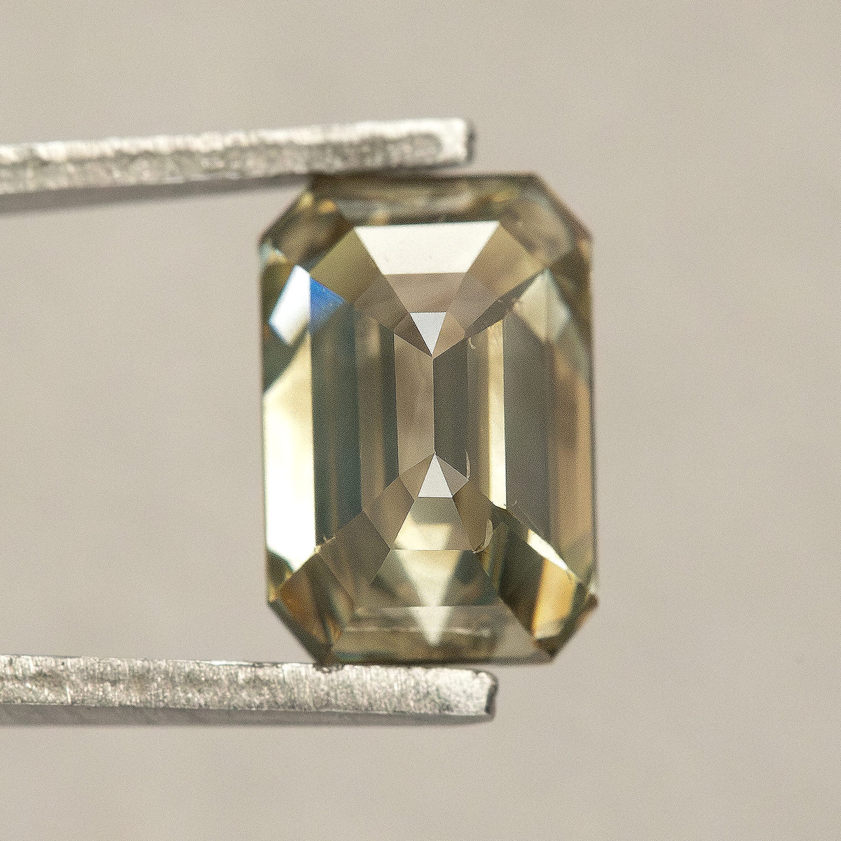 Reverse-facing emerald-cut diamond in tweezers, showing broad pavilion facets and a central lengthwise facet junction.