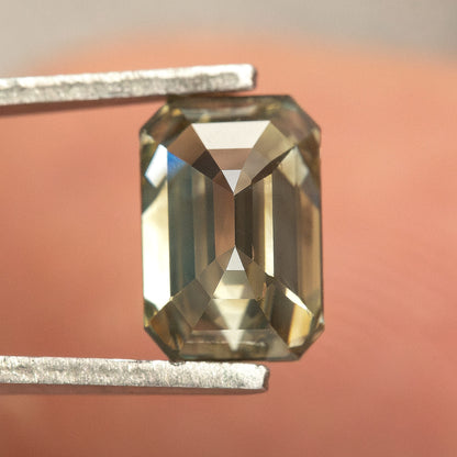 Pavilion view of the brown-green diamond in tweezers in front of a finger, with broad pale and dark facet reflections.