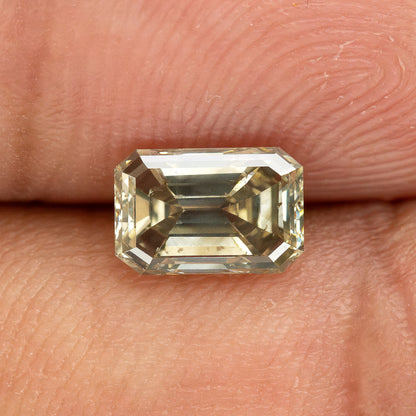 Emerald-cut diamond resting horizontally between fingertips, showing warm yellow-green reflections and skin ridges for scale.