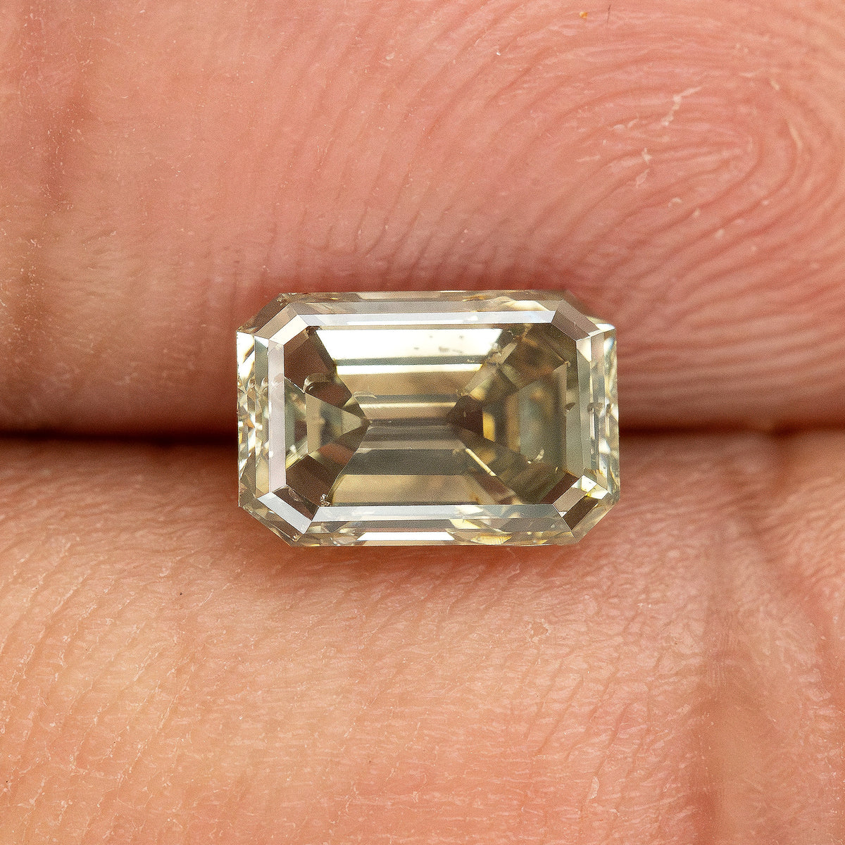 Horizontal fingertip view of the emerald-cut diamond at a changed angle, with pale step-facet reflections and darker internal areas.