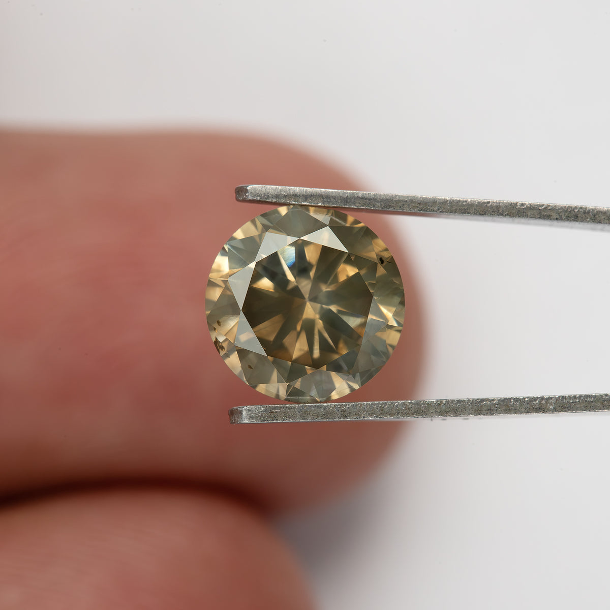KL444 held face-up in tweezers in front of a fingertip, showing the diamond at a smaller scale.