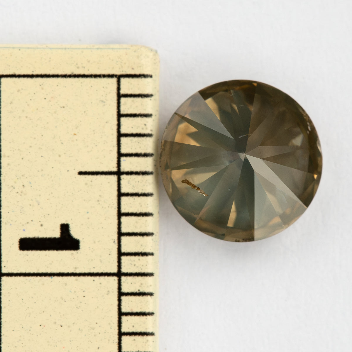 Pavilion-up view of the round diamond beside a millimeter ruler for size reference, showing olive-gray and champagne reflections and an elongated brown feature across one pavilion facet.