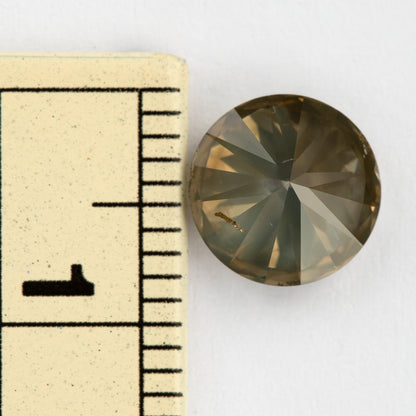 Pavilion-up view of the round diamond beside a millimeter ruler for size reference, showing olive-gray and champagne reflections and an elongated brown feature across one pavilion facet.