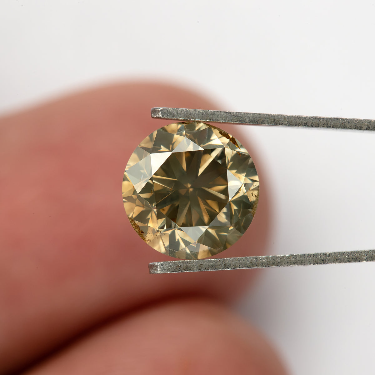 Face-up tweezers view with a fingertip behind the diamond for scale, showing darker olive-brown central reflections, pale-yellow outer facets and visible girdle features.
