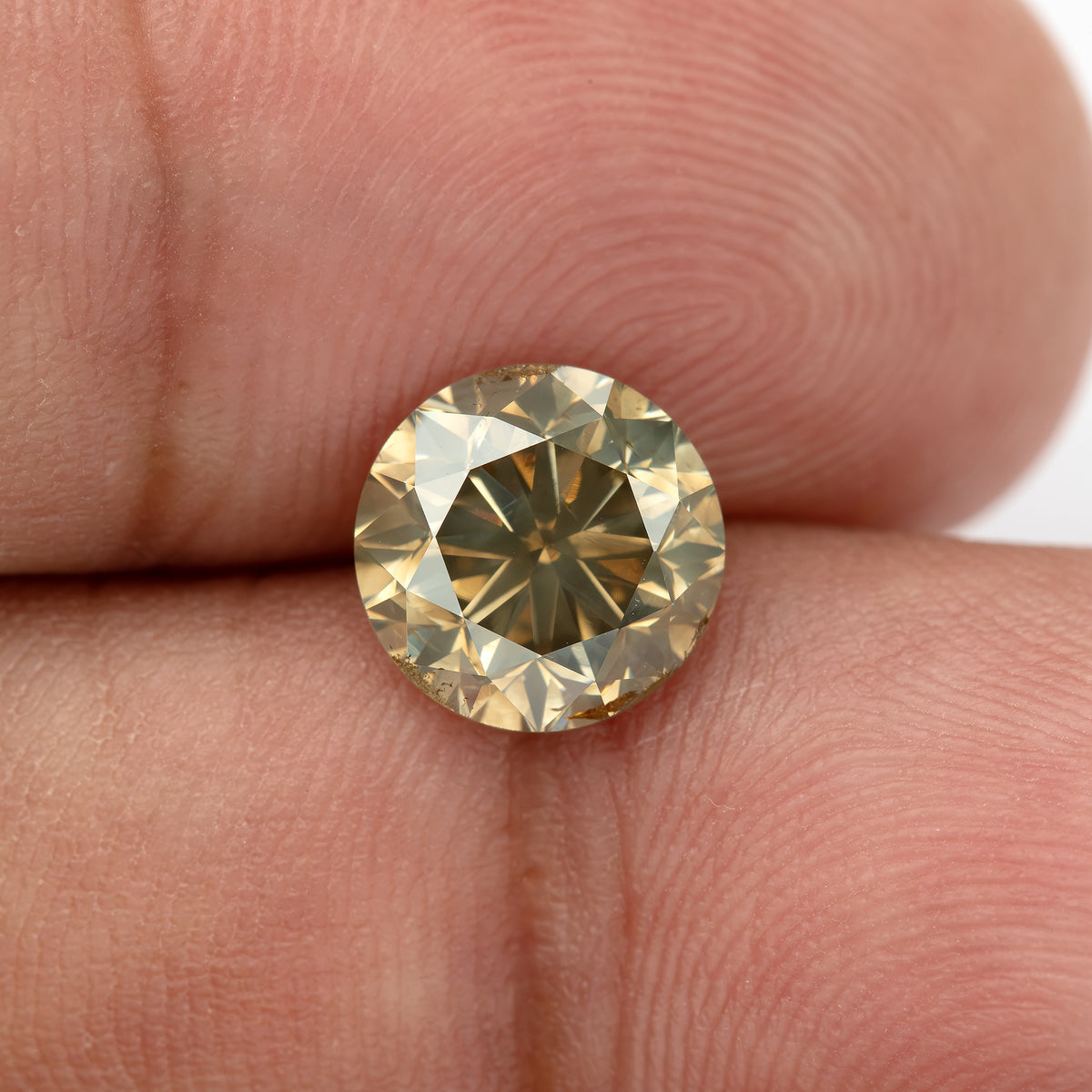 Close hand-scale view of the round brilliant diamond resting between fingers, showing its real-world size, olive-champagne color, pale-yellow flashes and visible brown girdle features.