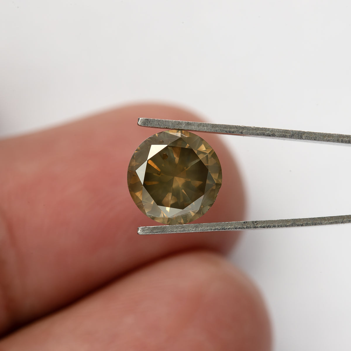 Diamante brillante redondo marrón oliva exacto sostenido con pinzas sobre la yema de un dedo para escala, con destellos dorados e inclusiones visibles debajo de la tabla.