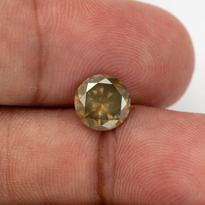 Vista cercana con escala de mano del diamante natural marrón oliva de 2.01 ct mostrando su contorno redondo, reflejos de facetas doradas y características de pureza visibles.