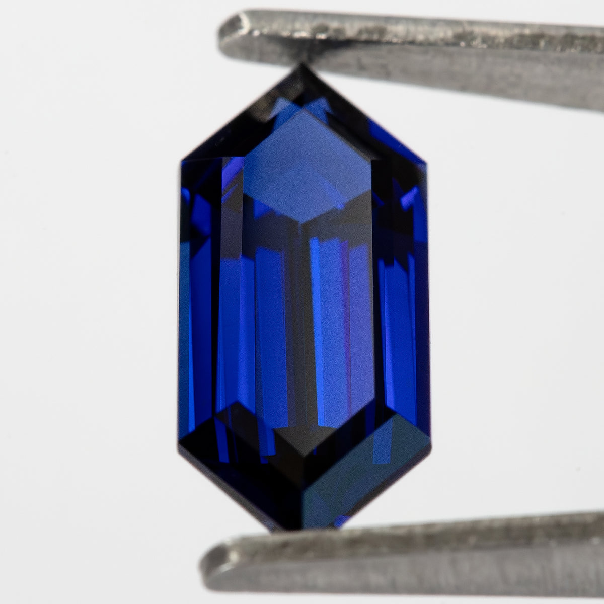 Tilted blue sapphire held vertically in tweezers, with bright reflections across its elongated face.
