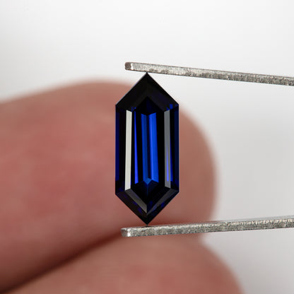 Blue elongated hexagonal sapphire held in tweezers in front of a fingertip for scale.