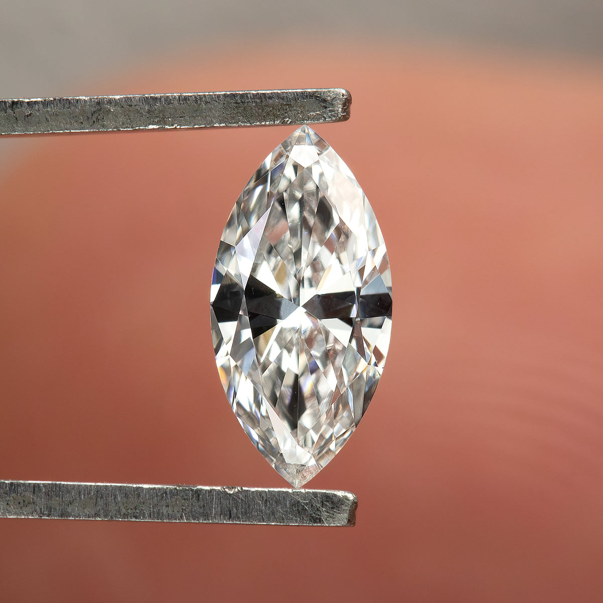 One marquise diamond held vertically face-up in tweezers in front of a finger, showing pointed ends and bright brilliant facets.