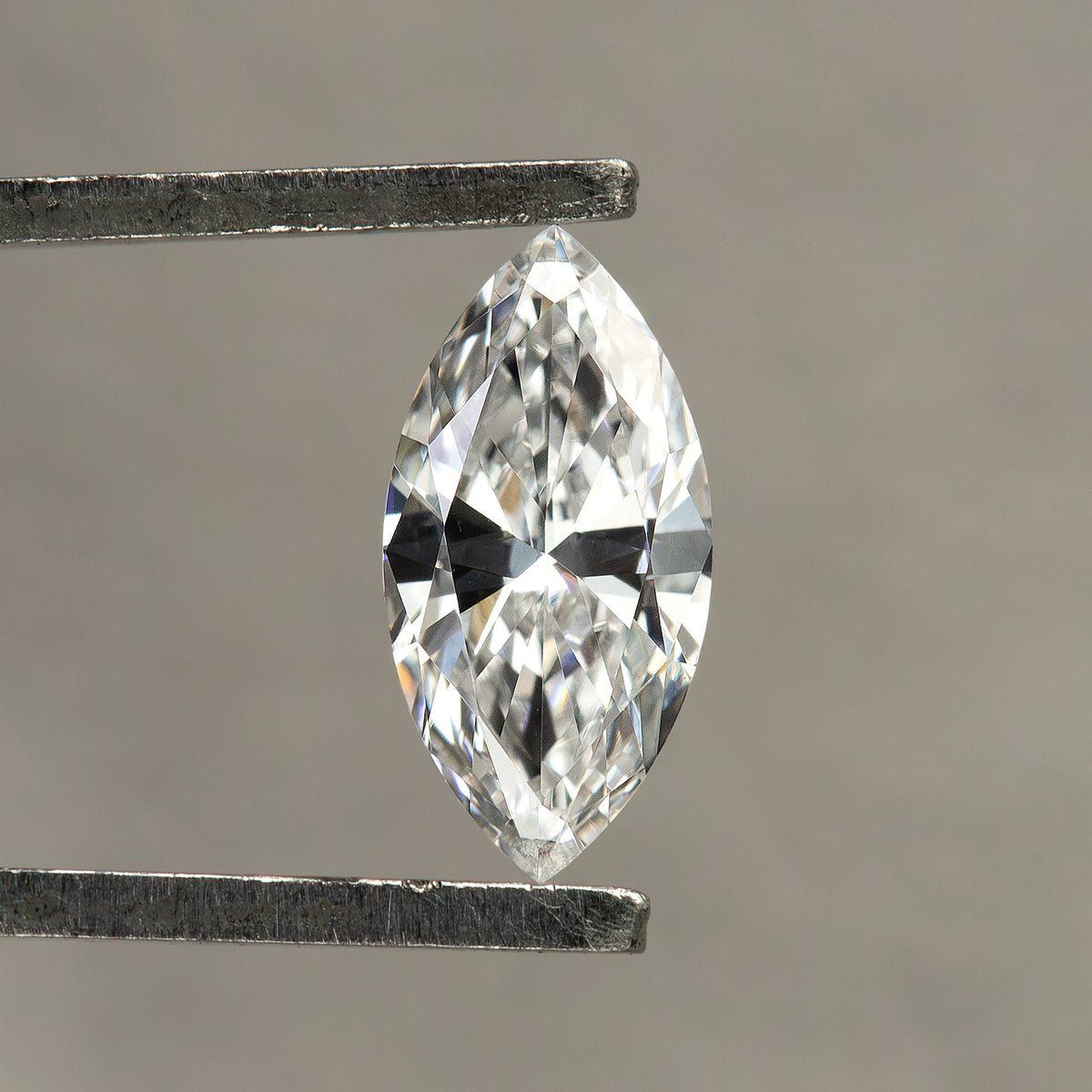 Single marquise diamond held face-up in tweezers against a neutral gray background, with white and dark reflections across its crown.