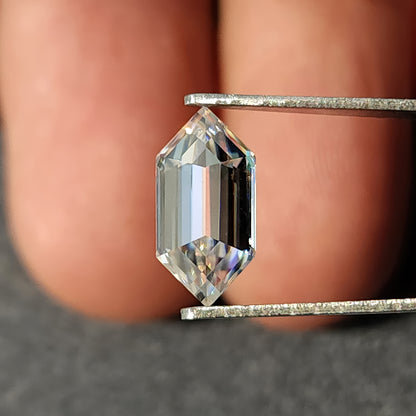 Vertical face-up view of an elongated hexagon step-cut transparent gem in tweezers held before fingers.