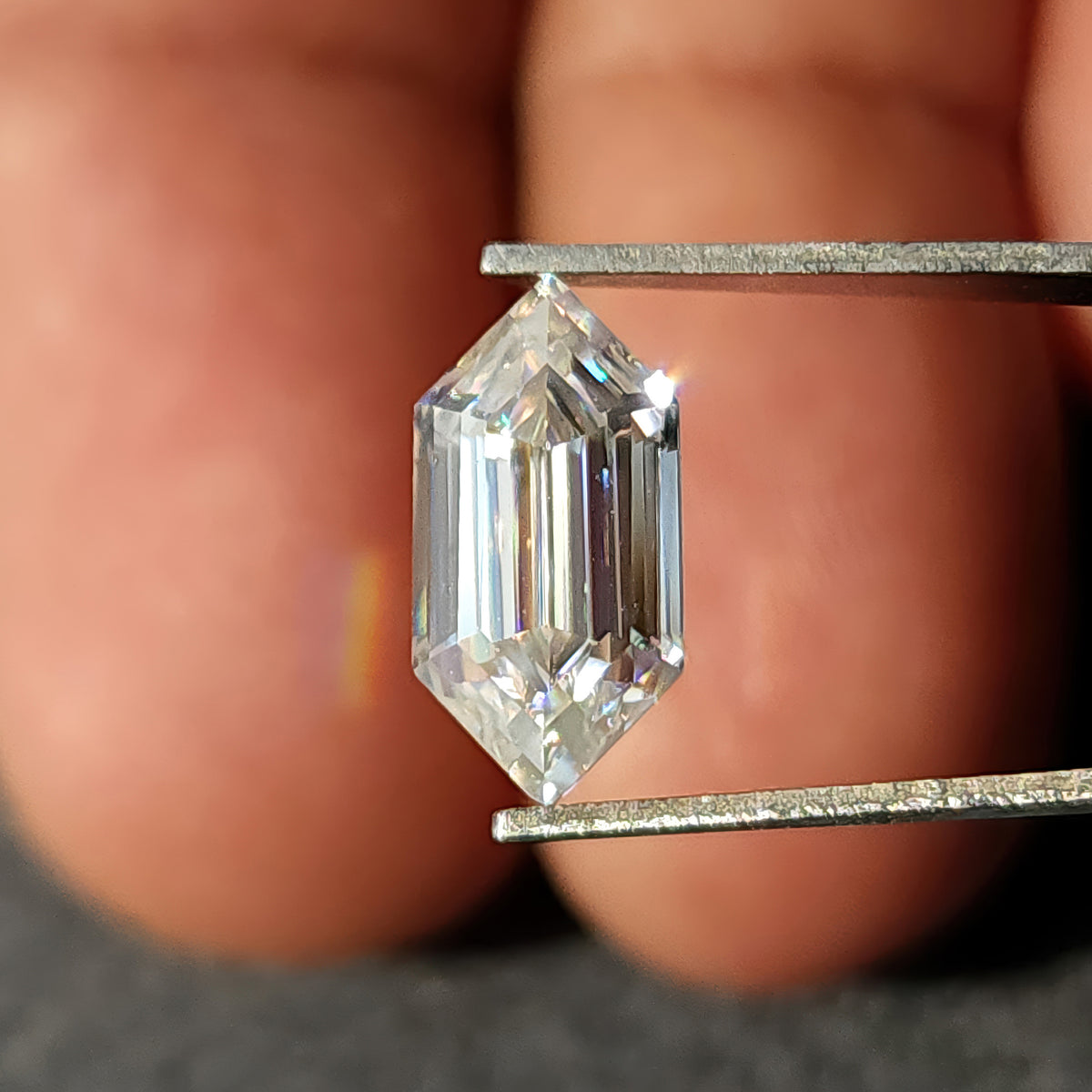 Elongated hexagon step-cut gem held in tweezers displaying strong fire and rainbow dispersion highlights.